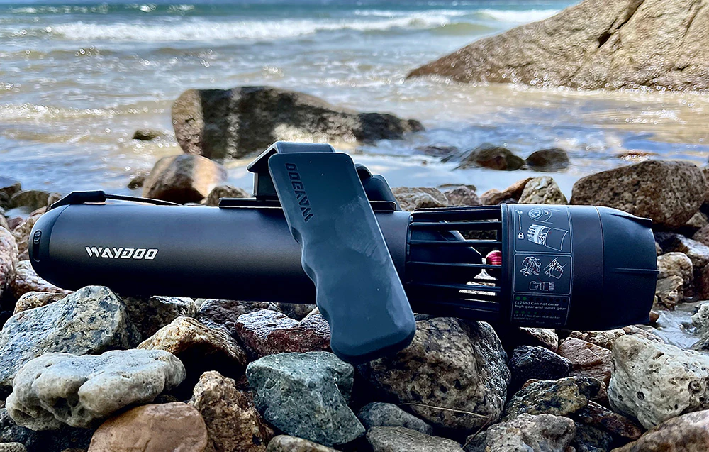 O unitate de propulsie electrică neagră pentru o placă de surf așezată pe un țărm stâncos, lângă apă.