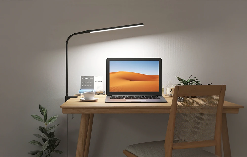 Un spațiu de lucru iluminat cu un laptop, cărți și o plantă. O lampă cu clemă este fixată pe partea laterală a unui birou din lemn.